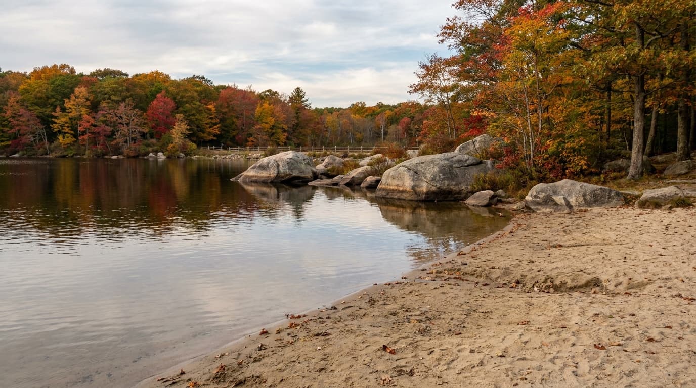 Calm Olney Pond at Lincoln Woods State Park ringed by autumn-colored woods and granite boulders, a freshwater swimming beach in the foreground, soft New England light, Rhode Island
