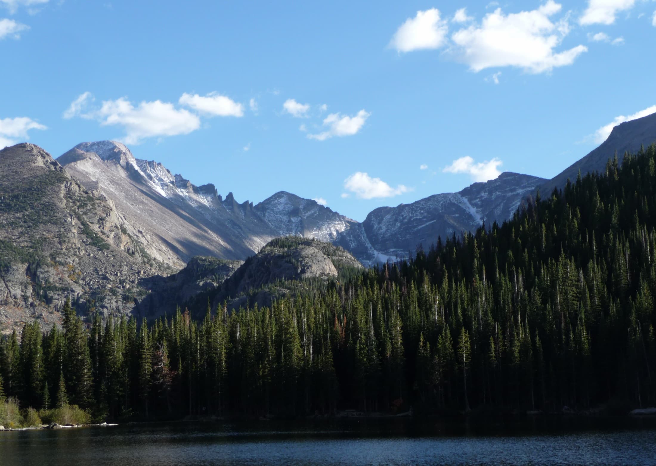 Glacier Gorge and rugged peaks rising above the still waters of Bear Lake, framed by pine forest, in Rocky Mountain National Park, Colorado