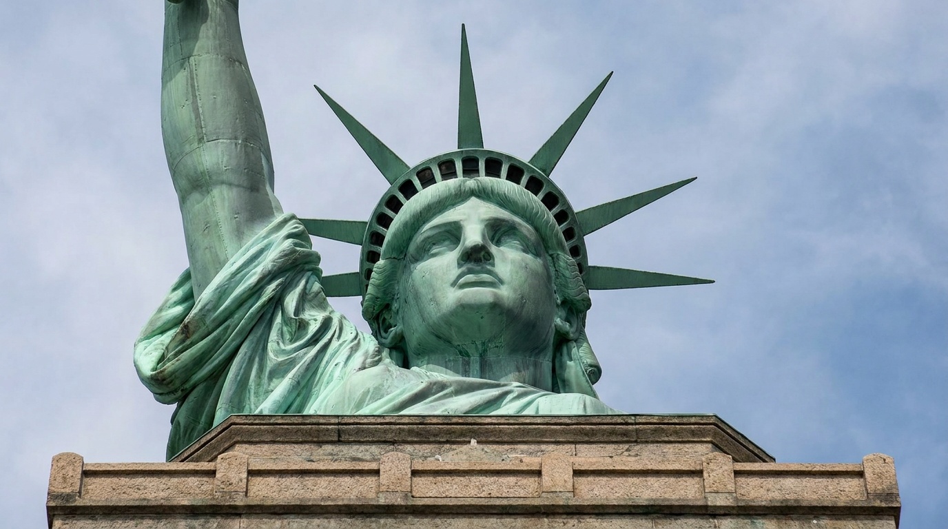 Looking up at the Statue of Liberty's crown and face from the pedestal level, copper detail and rivets visible against blue sky